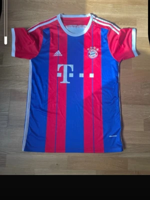 Bayern München matchtröja Adidas - Snygg Bayern München matchtröja från Adidas i rött och blått med vita detaljer. Klassiska ränder, klubbmärke och Adidas-logga på bröstet samt 'Bayern München' tryckt på ryggen. Tillverkad i lätt och ventilerande material, perfekt för fotbollsträning.