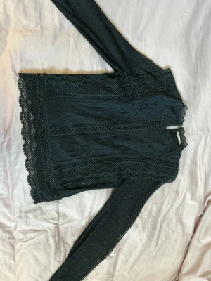 Svart spetsblus med lång ärm Only - Snygg svart topp i spets med långa ärmar och vågiga kanter. Transparenta partier på ärmarna och detaljerad spets framtill ger en elegant look. Perfekt för dig som gillar trendiga och feminina plagg.