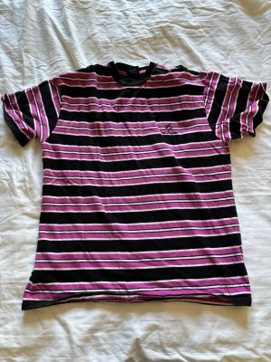 Randig t-shirt i svart och lila - Snygg randig t-shirt med svarta, lila och vita horisontella ränder. Klassisk passform med rund hals och korta ärmar. Perfekt för dig som gillar streetwear och vill sticka ut lite extra.