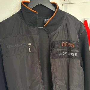 Svart vindjacka från Hugo Boss - Svart vindjacka från Hugo Boss med orange detaljer vid kragen och logga broderad på bröstet. Jackan har dragkedja framtill, en bröstficka med dragkedja och sportig känsla. Perfekt för dig som gillar stilrena och funktionella plagg. Hör av er för fler bilder.
