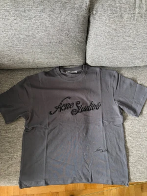  Acne Studios t-shirt - Säljer en grå t-shirt från Acne Studios med broderad logga i svart på bröstet och mindre logga nedtill. Klassisk rund hals och korta ärmar. Skön passform och stilren design, perfekt till jeans eller shorts.