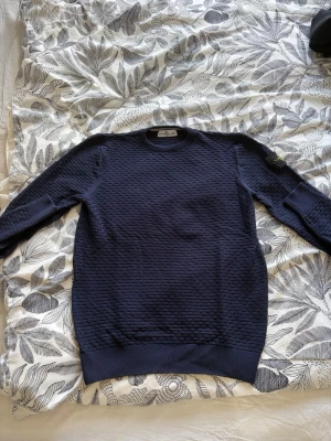 Mörkblå tröja från Stone Island - Säljer en mörkblå tröja från Stone Island med unik struktur och klassisk patch på ärmen. Tröjan har rund halsringning, ribbade muddar och är tillverkad i ett mjukt material som känns riktigt skönt. Perfekt för dig som gillar streetwear och snygga detaljer. Priset kan diskuteras!
