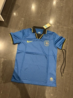 Blå AIK fotbollströja från Nike - Snygg blå AIK fotbollströja från Nike med svart krage och gula detaljer. Tröjan har AIK:s klubbmärke på bröstet, gul Nike-logga och är tillverkad i Dri-FIT-material som andas. Perfekt för träning eller match.