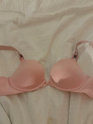 Ljusrosa push-up bh från Victoria's Secret - Säljer en ljusrosa push-up bh från Victoria's Secret med vadderade kupor och justerbara axelband. Bh:n har en slät design och axelbanden har märkets logga tryckt på insidan. Perfekt för dig som vill ha extra lyft och snygg passform.