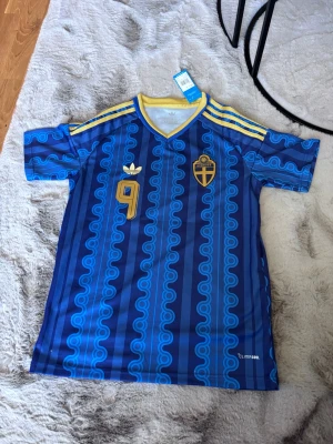 Blå Sverige fotbollströja Adidas - Säljer en blå Sverige fotbollströja från Adidas med gult nummer 9 och namnet Isak på ryggen. Tröjan har gula detaljer, klassiska Adidas-ränder på axlarna och Sveriges landslagsemblem på bröstet. Tillverkad i lätt och ventilerande climalite-material.