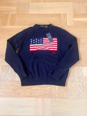 Mörkblå stickad tröja Polo Ralph Lauren - Mörkblå stickad tröja från Polo Ralph Lauren med amerikansk flagga i rött, vitt och blått på bröstet samt RL-broderi. Tröjan har rund halsringning och ribbade muddar vid ärmslut och nederkant. Perfekt för dig som gillar klassisk preppy stil.