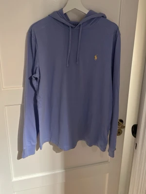 Ljuslila hoodie från Ralph Lauren - Snygg ljuslila hoodie från Ralph Lauren med klassisk huva och dragsko. Tröjan har lång ärm och den ikoniska gula loggan broderad på bröstet. Tillverkad i mjukt och lätt material som passar perfekt för en avslappnad stil.