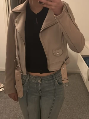 Beige/smutsrosa jacka från Zara - Snygg jacka från Zara med bred krage, silverfärgade dragkedjor och detaljer samt bälte i midjan. Jackan har en ficka med lock och en figurnära passform som ger en cool look. Perfekt för dig som vill ha en trendig och stilren jacka.