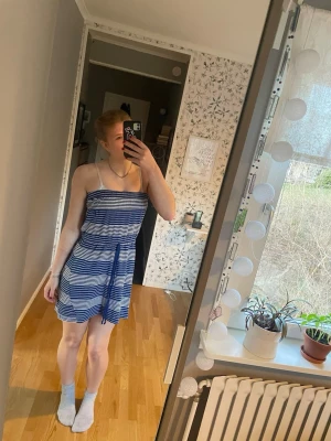 Blåvit randig kort sommarklänning - Säljer en blåvit randig kort klänning med knytband i midjan. Klänningen har en avslappnad passform och är perfekt för varma dagar. Tillverkad i ett mjukt och lätt material som känns skönt mot huden.