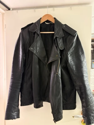 Svart klassisk skinnjacka från Whyred - Säljer en svart skinnjacka i bikerstil med breda slag, axelklaffar och sneddraget blixtlås. Jackan har detaljer som tryckknappar och dragkedjor vid ärmsluten. Perfekt för dig som vill ha en tidlös och cool look.