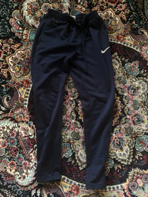 Mörkblå Nike mjukisbyxor - Snygga mörkblå mjukisbyxor från Nike med vit logga på låret. Byxorna har dragsko i midjan och sidofickor, samt smal passform som sitter skönt. Perfekta för chill eller träning.