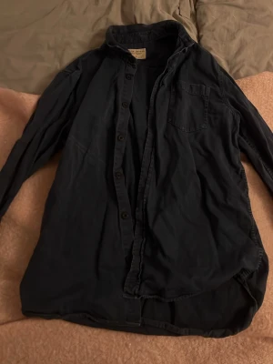 Mörkblå skjorta från Nudie Jeans Co - Säljer en mörkblå skjorta från Nudie Jeans Co i 100% ekologisk bomull. Skjortan har klassisk krage, bröstficka och knappar framtill. Perfekt för dig som gillar en enkel och stilren look. Lite urtvättad