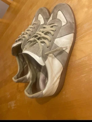 Maison Margiela Gats - !Intressekoll!Maison Margiela sneakers med vit läderbas och grå mockadetaljer. Klassisk gummisula i brunt och rund tå. Skorna har snörning och kommer med dustbags och originalkartong. Perfekta för dig som gillar stilrena och lyxiga sneakers.