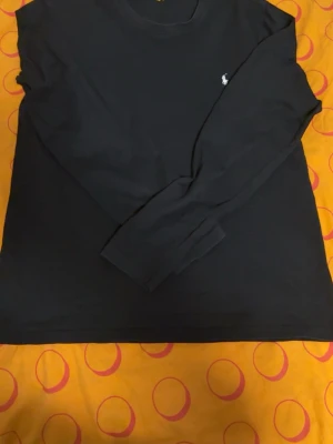 Svart långärmad tröja från Polo Ralph Lauren - Svart långärmad tröja från Polo Ralph Lauren med klassisk rund halsringning och den ikoniska vita broderade loggan på bröstet. Tröjan är i mjuk bomull och har en stilren, enkel design som passar till det mesta. Du kan ha den när som helst men perfekt nu under våren och sommaren sedan kan du använda den på vintern också 