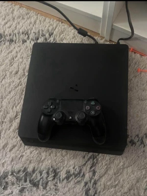 PlayStation 4 Slim med handkontroll - Säljer en svart PlayStation 4 Slim från Sony med tillhörande svart DualShock 4-handkontroll. Konsolen har en stilren, fyrkantig design i matt plast och är perfekt för gaming och streaming. Strömkabel medföljer.