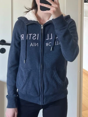 Blå zip hoodie från Hollister - Mysig blå hoodie från Hollister med dragkedja, vit fluffig foder på insidan av huvan och stora fickor framtill. Tryck i vitt med texten 'Hollister California' över bröstet. Perfekt för chill dagar och enkel att matcha med jeans eller leggings.