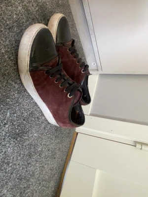 Vinröda sneakers med svart tå - Snygga vinröda sneakers med svart tåparti i blankt material och svarta skosnören. Skorna har en vit sula och är låga i modellen. Ovandelen verkar vara i mocka eller liknande material. Perfekt för dig som vill ha en unik look.