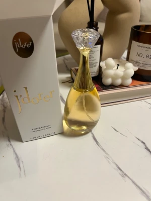 J'adore Eau de Parfum 90 ml - HELT FULL!!! Elegant parfymflaska från J'adore med en droppformad design och transparent glas. Flaskan har en guldtonad hals och en kristalliknande topp. Parfymen kommer i en vit kartong med guldtext och rymmer 90 ml Eau de Parfum.