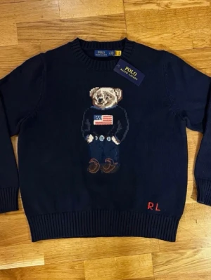 Mörkblå stickad tröja Polo Bear - Mörkblå stickad tröja från Polo Ralph Lauren med ikonisk Polo Bear-motiv på bröstet. Rund halsringning och ribbade muddar. RL-broderi i rött på nederkanten. Perfekt för dig som gillar klassisk och lekfull stil.