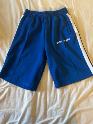 Blå Palm Angels shorts med vit stripe - Snygga blå shorts från Palm Angels med vit sidostripes och logga på vänster ben. Elastisk midja och två fickor framtill. Perfekta för sommaren och har en sportig vibe som passar till streetwear-stilen.