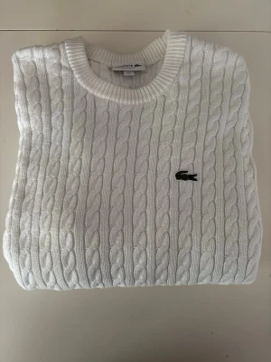 Vit kabelstickad tröja Lacoste - Vit kabelstickad tröja från Lacoste med rund halsringning. Tröjan har ribbade muddar och är gjord i mjukt material. Tröjan är i väldigt bra skick. Inköpt på Åhléns och kvitto finns. 