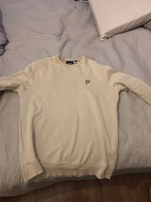 Beige sweatshirt från Lyle & Scott - Snygg beige sweatshirt från Lyle & Scott med klassisk gul logga på bröstet. Tröjan har rund halsringning, ribbade muddar vid ärmslut och nederkant samt en avslappnad passform. Perfekt för en stilren och enkel look.