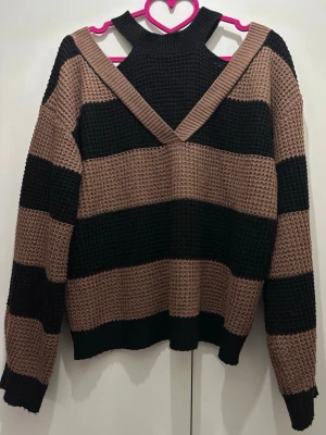 Randig stickad tröja med cut out-detalj - Stickad tröja med breda svarta och bruna ränder, långärmad och med unik cut out-detalj vid axlarna. Tröjan har en hög halsringning och avslappnad passform, perfekt för en trendig och bekväm look.