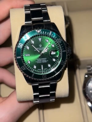 Rollie grön - Snygg Submariner med klassisk grön urtavla och vridbar bezel. Klockan har en robust länk i rostfritt stål och tydliga indexmarkeringar. Perfekt för dig som gillar tidlös design och exklusiv känsla. Automatisk rörelse och datumfönster vid 3-positionen.
