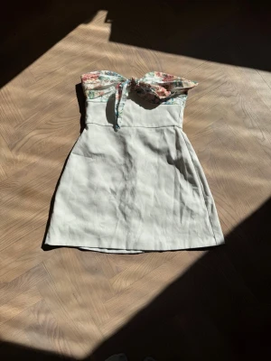 Vit kort klänning med blommig topp - Söt kort klänning med vit kjol och blommigt mönster upptill. Axelbandslös design och lätt utsvängd kjol. Perfekt för varma dagar och somriga tillfällen.