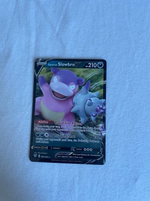 Pokémonkort Galarian Slowbro V - Säljer ett Pokémonkort med motivet Galarian Slowbro V. Kortet har färger i lila, rosa och grått med detaljerad illustration och text på engelska. Det är en V-kort med 210 HP och attacker som Rapid-Fire Poison och Tripping Shot. Perfekt för samlare eller Pokémonfans.