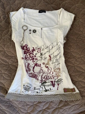 Vit topp med tryck - Vit topp med unikt tryck i vinrött och svart, dekorerad med text och blommor. Märket är TAIFUN collection. Det står inte någon storlek men jag skulle säga S-M. Pris går att diskuteras🤍