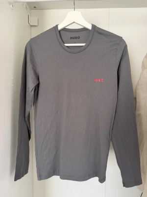 Grå långärmad topp från HUGO - Snygg grå långärmad topp från HUGO med rund halsringning och diskret röd logga på bröstet. Tillverkad i mjuk bomull som känns skön mot huden. Enkel och stilren design som funkar till det mesta.