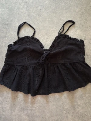 Svart linnetopp med spetsdetaljer - Snygg svart linnetopp med tunna axelband, volangkant och dekorativ snörning framtill. Toppen har fina spetsdetaljer och en lätt rynkad nederdel som ger en feminin look. Perfekt att matcha med jeans eller kjol.