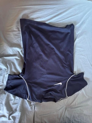 Svart Nike sporttröja med vita detaljer - Svart sporttröja från Nike med korta ärmar och vita kontrastsömmar längs sidorna. Tröjan har en klassisk Nike-logga på bröstet och är tillverkad i ett lätt och snabbtorkande material, perfekt för träning och sportaktiviteter. Strl 147-158cm