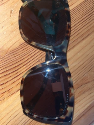 Ralph Lauren solglasögon leopardmönster - Snygga solglasögon från Ralph Lauren med leopardmönstrad båge och mörka glas. Modellen har breda skalmar med silverfärgade RL-detaljer på sidorna. Perfekt accessoar för att lyfta din stil under soliga dagar.