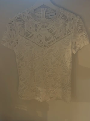 Vit spetsblus från VILA - Säljer en superfin vit spetsblus från VILA med korta ärmar och rund hals. Blusen har ett detaljerat spetsmönster och är lätt transparent, perfekt för att skapa en chic look. Passar till både jeans och kjol för en trendig outfit.