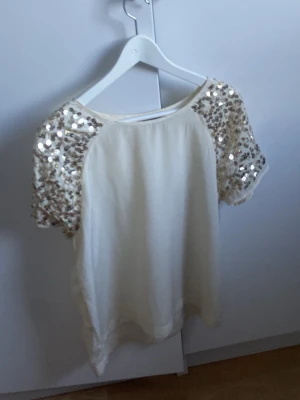 Vit topp med paljettärmar - Snygg vit topp med korta ärmar täckta av glittriga paljetter i guld. Rak passform och rund halsringning. Perfekt för dig som vill sticka ut lite extra. Materialet känns mjukt och lätt, troligen polyester.