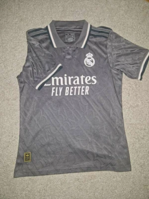Real Madrid grå matchtröja Adidas - Snygg grå Real Madrid matchtröja från Adidas med vita detaljer och klubbmärke på bröstet. Tröjan har korta ärmar, krage och tryck med 'Emirates Fly Better' på framsidan. Perfekt för fotbollsträning eller att bära som supporter. Det står M på lappen, men den är liten i storleken. Den passar bäst för dig som vanligtvis bär S eller till och med XS. Passar bäst för någon som är ca 160–170 cm lång