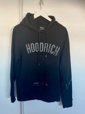Svart hoodie från Hoodrich - Svart hoodie från Hoodrich med stor logga på bröstet och tryck på ryggen. Hoodien har huva med dragsko, lång ärm och en stor magficka. Materialet är mjukt och bekvämt, perfekt för en avslappnad streetstyle-look.