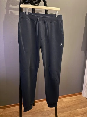 Svarta mjukisbyxor från Polo Ralph Lauren - Svarta mjukisbyxor från Polo Ralph Lauren med dragsko i midjan och ribbade muddar vid bensluten. Byxorna har sidofickor och den klassiska vita Polo-loggan broderad på vänster ben. Perfekta för en avslappnad och stilren look.