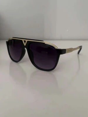 Svarta solglasögon - Snygga svarta solglasögon med guldiga detaljer och mörka glas. Modellen har en markerad ram och klassisk pilotform med en tydlig detalj i metall upptill. Perfekt accessoar för att lyfta din stil.