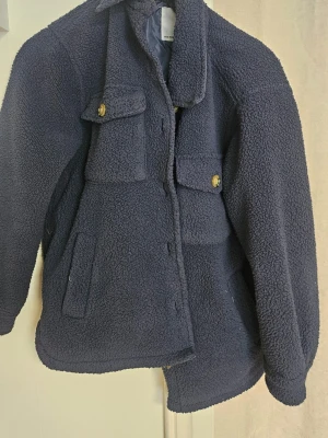 Mörkblå teddyjacka med fickor - Mysig mörkblå teddyjacka med två stora bröstfickor med knappar och två sidofickor. Jackan har klassisk krage och stängs med knappar framtill. Perfekt för kyliga dagar och ger en trendig look.