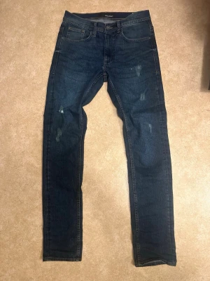 Mörkblå jeans från Blend - Snygga mörkblå jeans 29/32 från Blend med smal passform och slitna detaljer på benen. Klassisk femficksmodell med normal midja och dragkedjegylf. Perfekta för en avslappnad och trendig look.