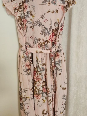 Ljusrosa blommig midiklänning - Söt ljusrosa midiklänning med blommigt mönster i beige, grått och vinrött. Klänningen har korta ärmar, markerad midja med avtagbart knytband och mjukt, lätt tyg som faller fint. Perfekt för dig som gillar romantisk och feminin stil.