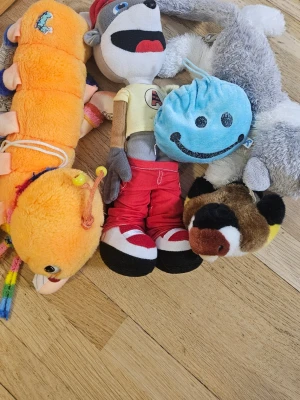 Blandade gosedjur och mjukisdjur - Säljer ett gäng blandade gosedjur: en lång orange tusenfoting med regnbågsfärgade ben, en apa med röda byxor och keps, en blå rund figur med smiley, en grå-vit kanin och en brun-vit fågel. Perfekt för dig som gillar mjukisdjur i olika färger och former.