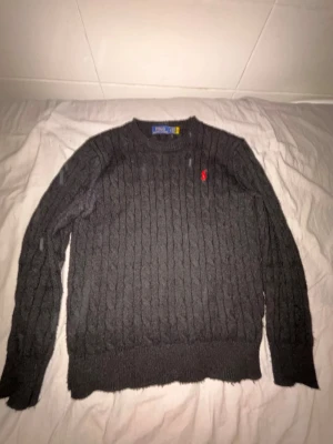 Mörkgrå stickad tröja Polo Ralph Lauren - Mörkgrå stickad tröja från Polo Ralph Lauren med klassiskt kabelstickat mönster och rund halsringning. Tröjan har långa ärmar och en liten röd broderad logotyp på bröstet. Perfekt för att hålla sig varm och snygg under kyliga dagar.