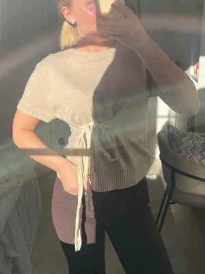 Beige stickad topp med knytband🩵 - Trendig beige stickad topp med korta ärmar och rund hals. Toppen har snygga knytband i midjan som ger en cool detalj och markerar figuren. Ribbstickad kant nedtill och vid ärmslut. Perfekt till både jeans och kjol för en avslappnad look.