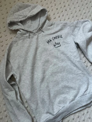 Grå hoodie med tryck 'Ma Chérie' - Ljusgrå hoodie med huva och dragsko. På bröstet och ryggen finns ett svart tryck med texten 'MA CHÉRIE' och en krona. Hoodien har ribbade muddar vid ärmslut och nederkant. Perfekt för en chill och avslappnad stil. OBS. Sömmen har gått upp lite vid “halsen”, se bild. 