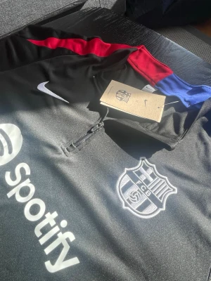 Svart FC Barcelona träningströja Nike - Svart långärmad träningströja från Nike med FC Barcelona-logga och Spotify-tryck på bröstet. Tröjan har dragkedja vid halsen och detaljer i rött och blått längs axlarna. Perfekt för fotbollsträning eller som supporterplagg.             Storlek: M 📏                                                          Frakt: Snabb och billig frakt 🚚 💴 (pris gå att diskutera). 