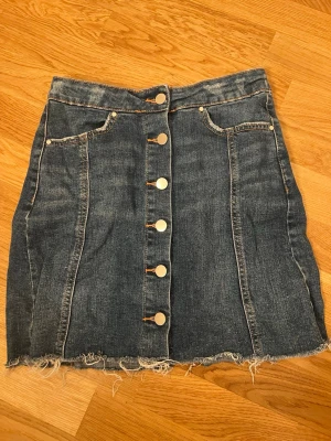 Jeanskjol med knappar från g perfect jeans - Säljer en blå jeanskjol från Gina trixig  jeans i storlek 34. Kjolen har knappar framtill, rå kant nertill och klassiska fickor fram. Perfekt för dig som gillar en avslappnad och trendig stil.